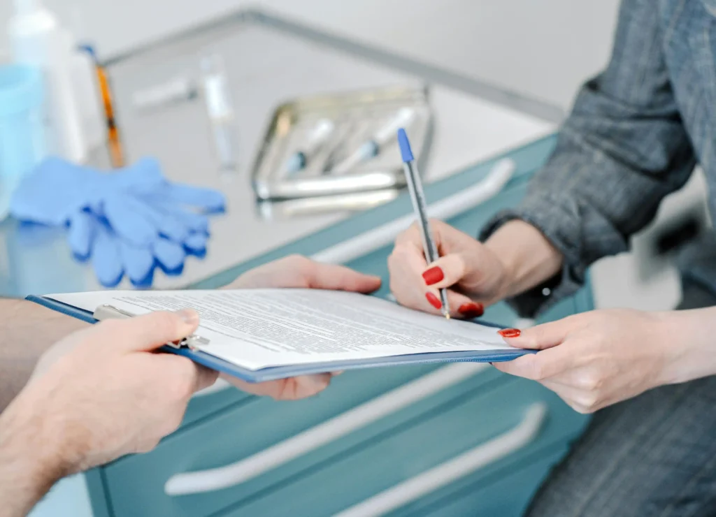 クリップボードに固定された書類にペンで署名をする様子。背景には医療用グローブや器具が置かれた、専門的・臨床的な環境。